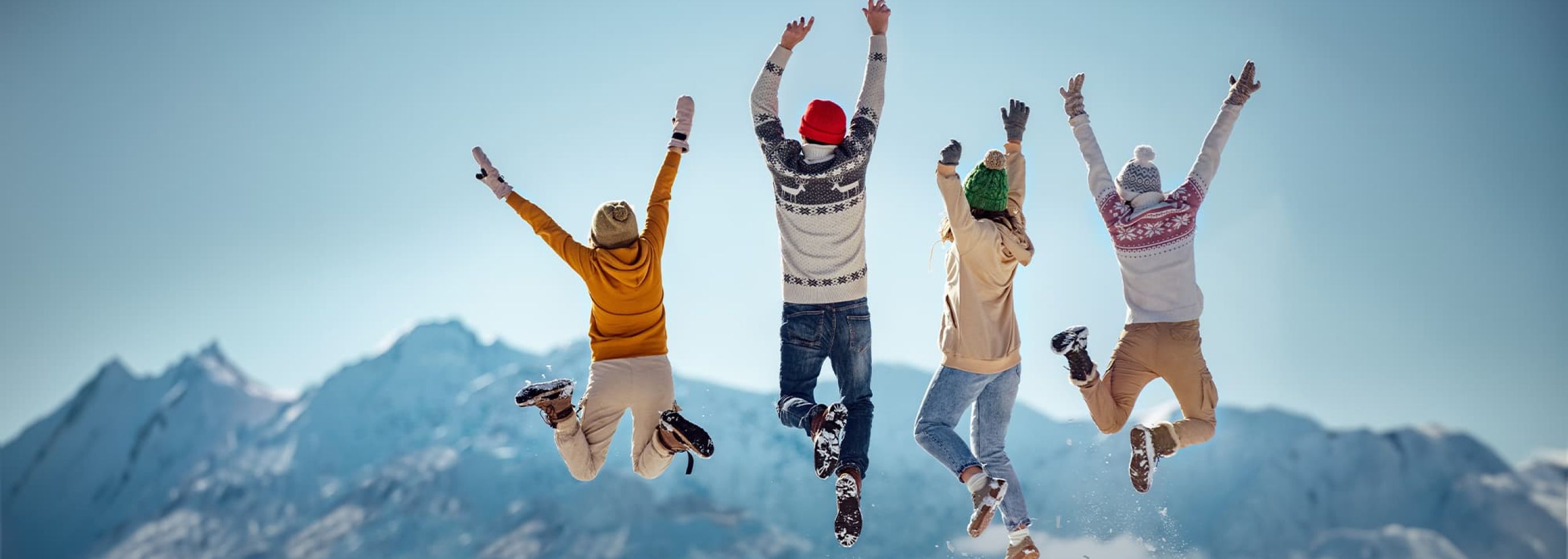Four enthusiastic students in winter clothing jump for joy with hands raised, facing away, against the backdrop of a stunning mountain range.