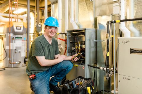 A male student wearing a helmet and safety goggles works on plumbing and piping equipment.
