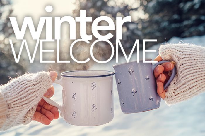 Winter Welcome text overlaid on a wintery scene with two gloved hands clinking mugs with warm beverages