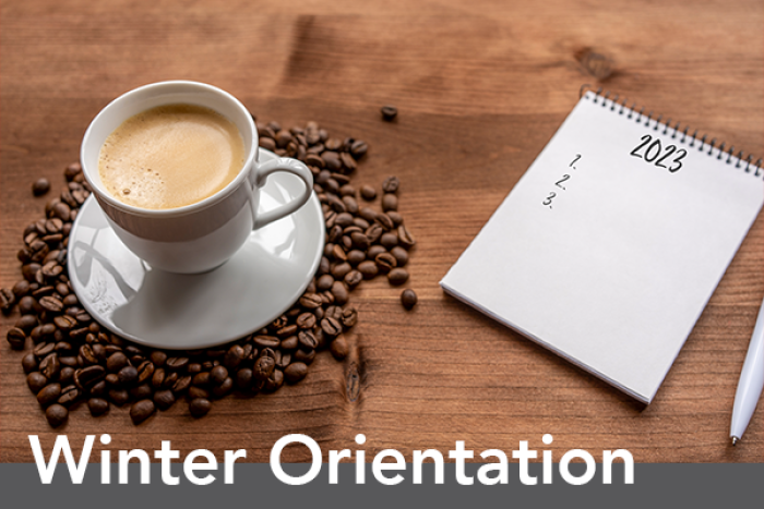 A white cup of frothy coffee sitting on a bed of dark brown coffee beans on a rustic wooden table. Beside the coffee is a white notepad with 2023 and 1, 2, 3 listed under with a white pen laying beside it. The words winter orientation are underneath