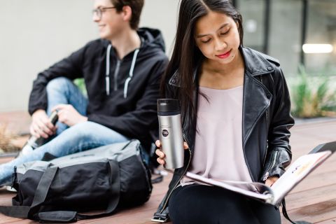 Female student studying outside and golding a thermos with a male student in the distance having a conversation.
