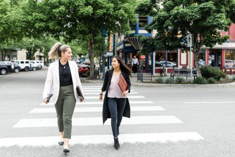Two female students walking in a crosswalk, having a conversation and carrying their books.