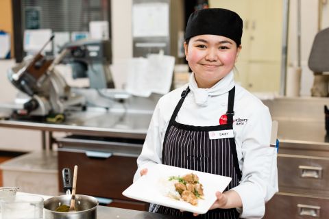 A female international student wearing an apron and chef's coat holds a plate of food. 