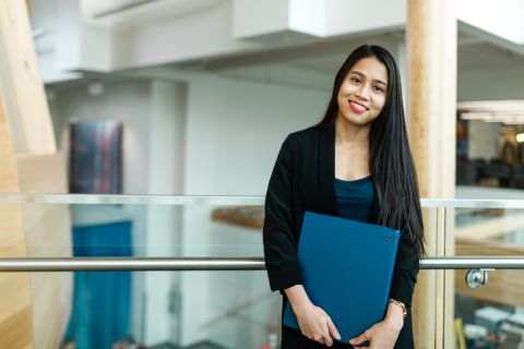 A female international student wearing a black blazer and red lipstick stands in a hallway holding a blue binder.