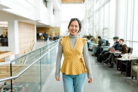 An international female student wearing a yellow top walking through a hallway of students studying.