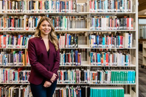 Female student standing in front of a shelf of books in the library wearing a blazer.