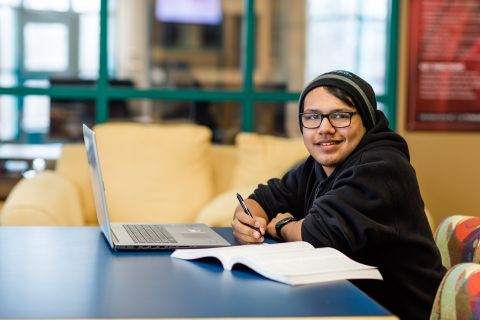 Male Indigenous student wearing glasses and a toque sits in the library studying.