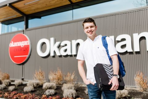 Male student wearing a short sleeve shirt carrying a backpack and books stands in front of the Okanagan College sign.