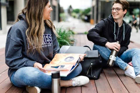 A female and male sit on a deck having a conversation  with their textbooks open.