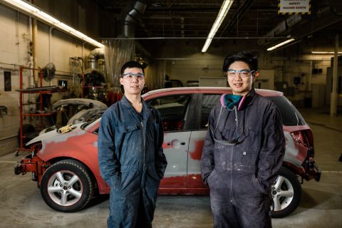Two Motor Vehicle Body Repairer Apprenticeship students stand in front of a vehicle in need of repair