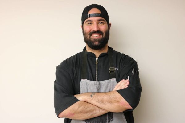 Alumnus Adam Relvas stands in front of a wall wearing a backwards hat and chef's coat.
