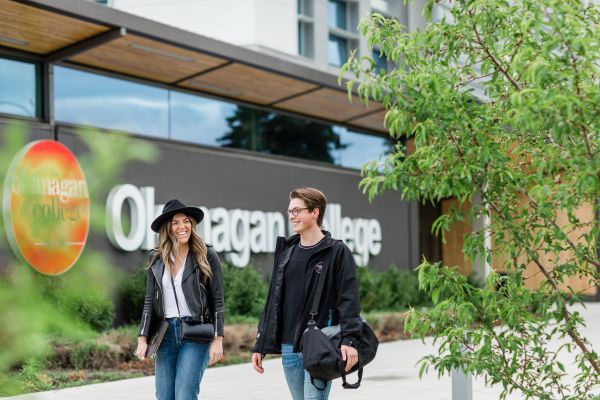 One female and one male business student walking outside in front of the Okanagan College sign.