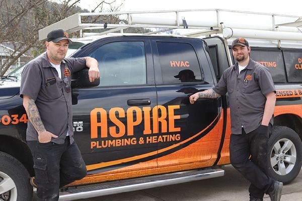 two males stand in front of a company truck with "Aspire Plumbing and Heating" printed on the outside