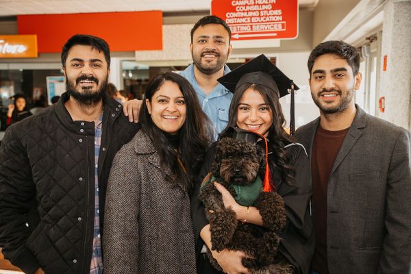 Graduate in cap and gown holding a dog wearing a graduation cap along with family members