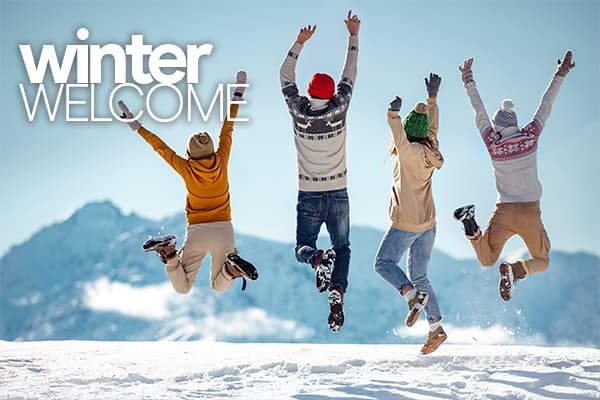 Four enthusiastic students in winter clothing jump for joy with hands raised, facing away, against the backdrop of a stunning mountain range. The accompanying text in the image reads, "Winter Welcome"
