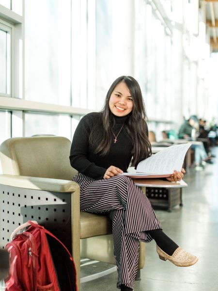 Smiling Okanagan College student in black shirt and striped pants sitting in a chair in the Centre for Learning Atrium holding a notebook with a backpack sitting at their feet.
