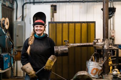 Female Welding student stands in front of equipment with her mask flipped open on her head.