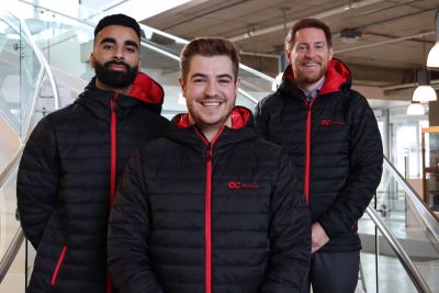 Three OC Finance team, business competitors, stand smiling wearing black and red Okanagan College jackets
