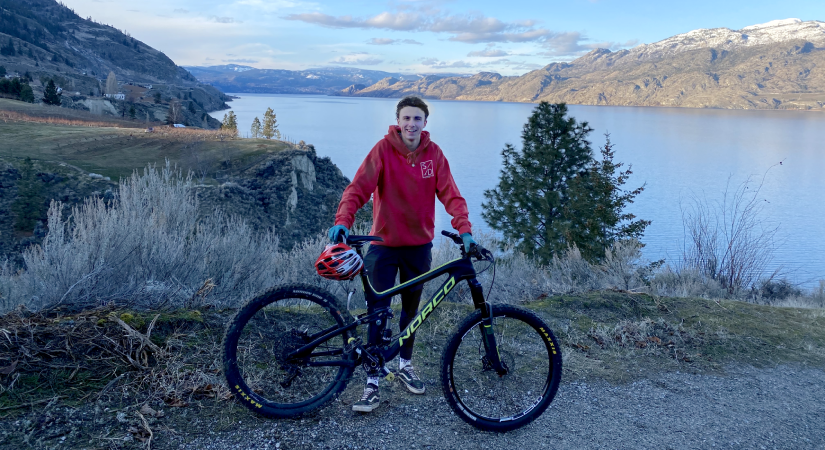 Kinesiology alumnus Ty Sideroff stands with his mountain bike with Skaha Lake in the view behind.