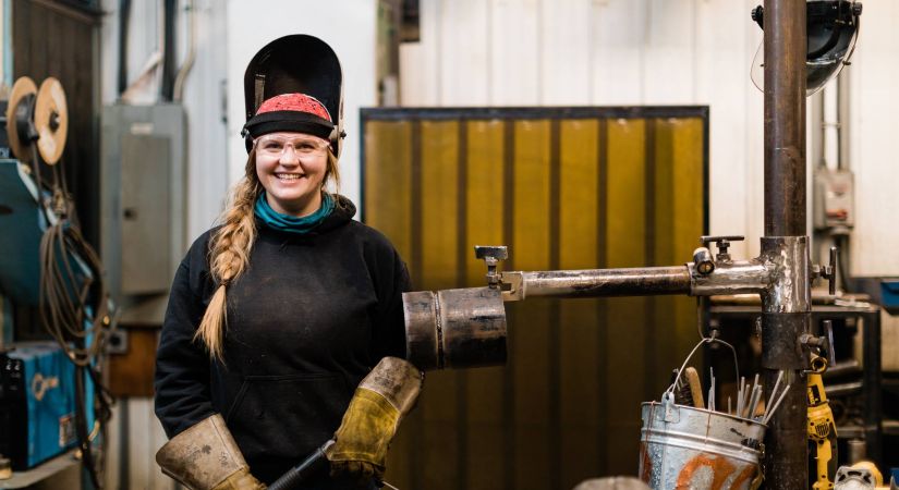 Female Welding student stands in front of equipment with her mask flipped open on her head.